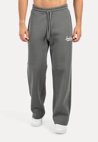 Pantalones de chándal gris claro, hechos de un tejido suave, con cintura ajustable y bolsillos laterales. Decorados con un logotipo de texto blanco en el muslo izquierdo.