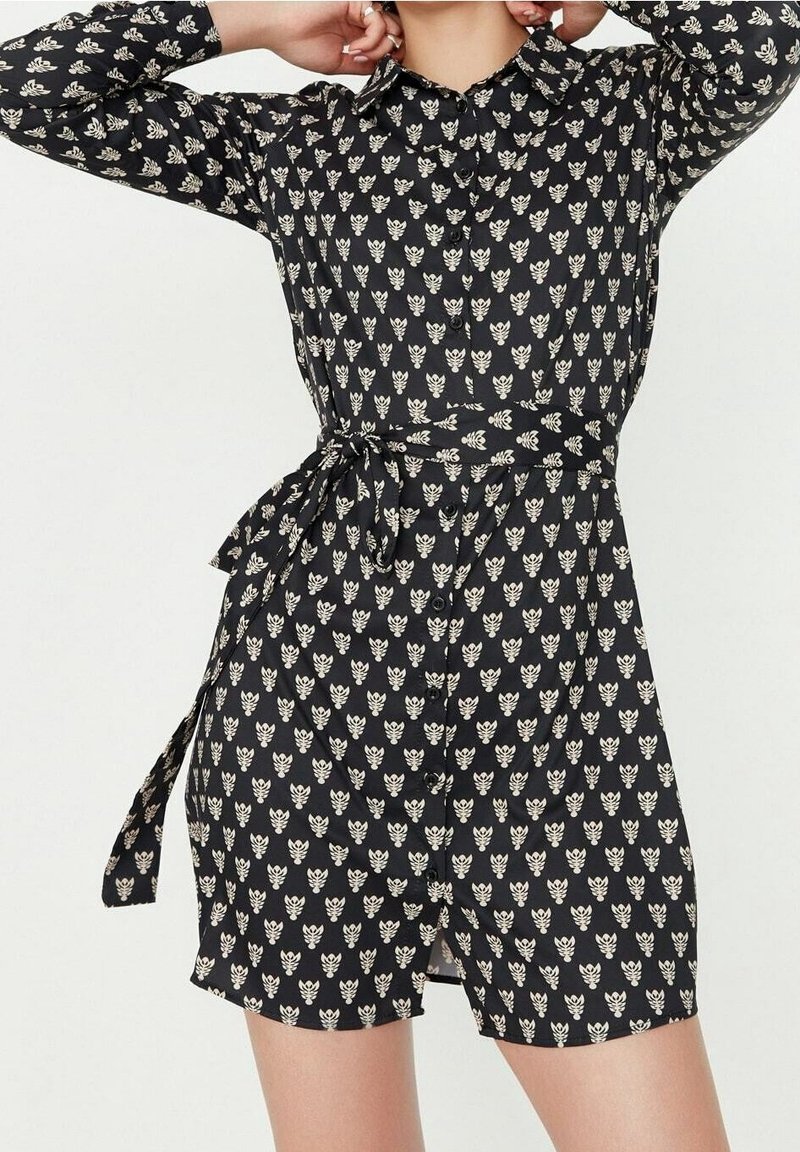 Robe chemise noire avec un motif floral crème, manches longues, col, boutonnage sur le devant et détail de taille nouée. Texture de tissu lisse.