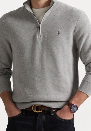 Homme portant un pull gris clair à fermeture éclair quart de zip, un jean foncé avec une ceinture marron et une montre-bracelet à cadran bleu avec un bracelet en cuir marron.