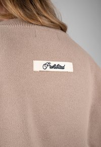 Personne blonde portant un pull en tricot beige avec une étiquette blanche inscrivant "Prohibited" cousue dans le haut du dos.