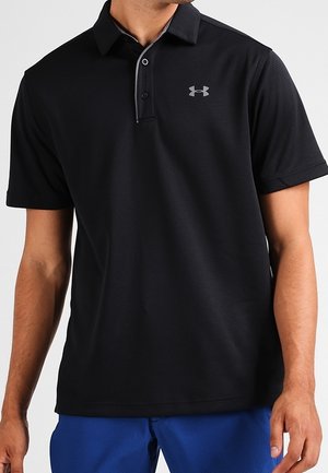 Homme portant un polo noir à manches courtes avec deux boutons et un logo gris Under Armour sur la poitrine, associé à un short bleu.