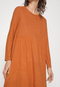 Vestido de color óxido, de mangas largas y escote redondo, con falda acampanada y detalle cosido en la cintura, hecho de una suave tela texturada.