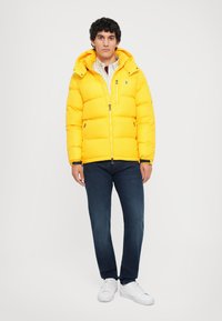 Man standing wearing a bright yellow puffer jacket, dark blue jeans, white sneakers, and a light-colored shirt against a plain background.