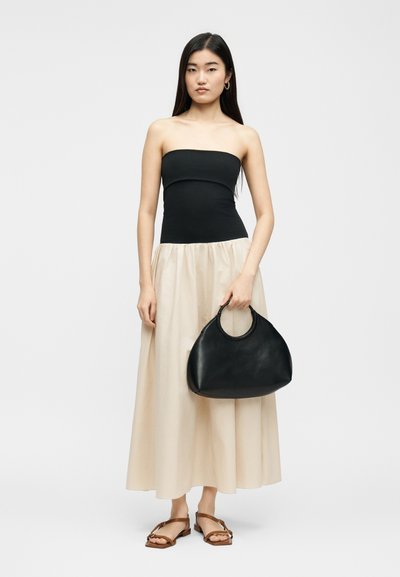 Γυναίκα που φορά ένα μαύρο τοπ χωρίς τιράντες και μια μπεζ φούστα, κρατώντας μια μεγάλη μαύρη τσάντα και φορά καφέ σανδάλια, στέκεται μπροστά σε απλό φόντο.
