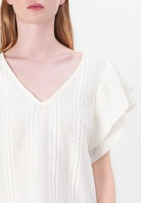 Blusa bianca con scollatura a V, tessuto texturizzato, maniche corte a volant e dettagli ondulati cuciti sulla parte anteriore.