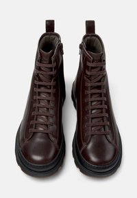 Botas de tobillo de cuero marrón con cordones en la parte delantera, cuello acolchado y suelas de goma. Presentan un cierre de cremallera y una lengüeta para una puesta fácil.
