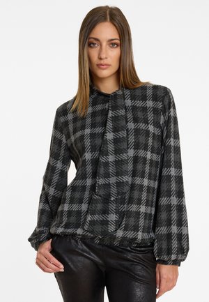 Blouse à carreaux noirs et gris avec une texture douce, présentant un col haut et des manches amples, associée à un pantalon noir brillant.