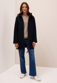 Zwarte fleecejas met een rits aan de voorkant, zijzakken en een capuchon, gedragen over een beige top en blauwe flared jeans, gecombineerd met witte sneakers.