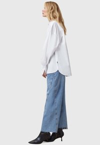 Weißes Langarmhemd mit lässigem Schnitt, seitlichen Schlitzen, kombiniert mit hellblauen weit geschnittenen Jeans und schwarzen spitzen Pumps.