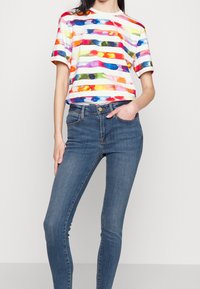 Camiseta de manga corta de rayas multicolores en algodón, combinada con jeans ajustados azules. La camiseta presenta patrones de acuarela llamativos sobre una base blanca.