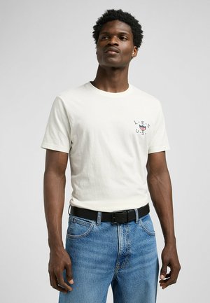 T-shirt en coton blanc à manches courtes et col rond, avec un petit graphique rouge et bleu sur la poitrine, associé à un jean bleu et une ceinture noire.