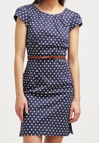 Marinblå klänning med vita prickar, korta ärmar, plisserad halsringning, figursydd midja och ett brunt bälte. Slät tyg med en lätt lyster.