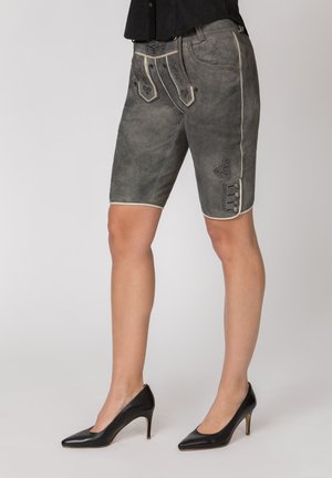 Frau trägt graue traditionelle Lederhosen mit Stickerei, schwarze spitze High Heels und ein schwarzes Oberteil, steht vor einfachem Hintergrund.