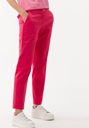 Henkilö, jolla on kirkkaan vaaleanpunaiset suorat housut taskuilla ja valkoiset korolliset tennarit, seisoo yksinkertaisen valkoisen taustan edessä.