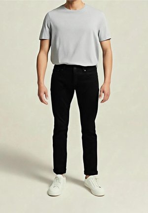 Homme portant une chemise à manches courtes gris clair unie, un pantalon noir slim et des baskets blanches, debout devant un fond neutre.