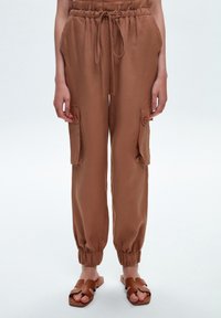 Pantalon cargo marron, ample, avec une taille élastique, des poches latérales et des poignets élastiques aux chevilles. Fabriqué en tissu léger.