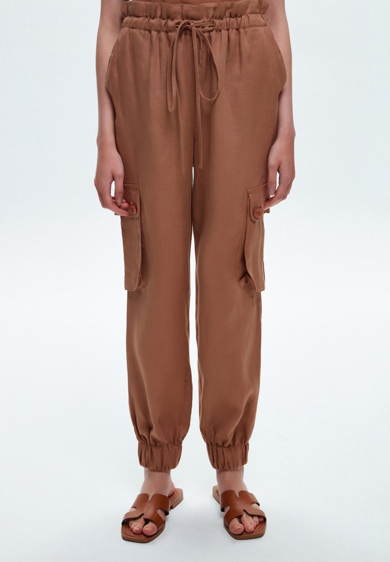 Pantalon cargo marron, ample, avec une taille élastique, des poches latérales et des poignets élastiques aux chevilles. Fabriqué en tissu léger.