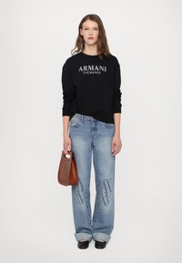 Zwarte sweatshirt met het logo "ARMANI EXCHANGE", lichtblauwe jeans met logo-accenten, bruine handtas, zwarte schoenen, lang bruin haar.