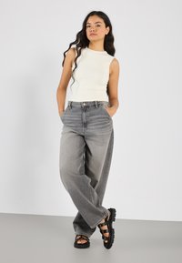 Top senza maniche color crema, jeans in denim grigi ad alta vita con vestibilità ampia e sandali neri con dettagli sulle cinghie, su sfondo neutro.