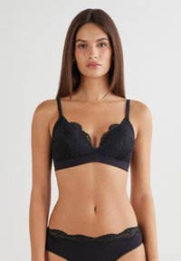 Svart spets-bralette med blommönster, djup urringning, justerbara band och en mjuk resår. Designad med en delikat vågig kant.