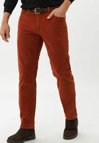 Uomo che indossa pantaloni in corduroy color ruggine, camicia nera, cintura marrone e stivali in suede marrone scuro, in piedi con una mano in tasca.