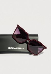 Bordó négyzet alakú napszemüveg sötét lencsékkel, fekete tokra téve, amelyen fehér betűkkel a "KARL LAGERFELD" felirat olvasható.
