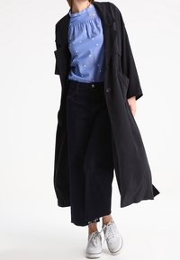 Manteau long noir sur un chemisier bleu clair avec des motifs d'étoiles blanches, associé à un jean court foncé et des baskets blanches.