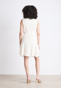 Sleeveless dress in light beige with vertical pleats and a fitted waist. Flared skirt with a textured, chevron pattern. Back view.