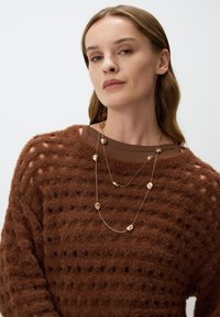 Bruine fuzzy trui met een gestructureerd patroon, gelaagd gouden collier met onregelmatig gevormde accenten, gedragen door een model tegen een lichte achtergrond.