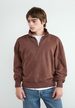 Jeune homme portant un sweat-shirt marron à fermeture éclair sur une chemise blanche avec un jean bleu, regardant vers la gauche sur un fond clair uni.