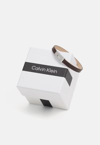 Bracciale in pelle marrone con chiusura in metallo argento, esposto su una scatola regalo bianca Calvin Klein con una striscia nera.