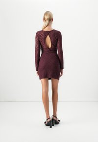 Femme avec une queue de cheval blonde portant une robe en dentelle bordeaux ajustée à manches longues avec une découpe dans le dos et des sandales à talons hauts noires, se tenant sur un sol blanc.