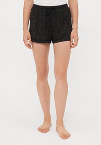 Kvinna som bär svarta och mörkbruna leopardmönstrade shorts med dragsko, stående barfota mot en vit bakgrund.