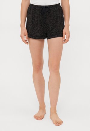 Kvinna som bär svarta och mörkbruna leopardmönstrade shorts med dragsko, stående barfota mot en vit bakgrund.