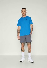 Mann med tatoveringer iført en blåmønstrete t-skjorte, grå shorts, hvite sokker og hvite joggesko som står mot en enkel lys bakgrunn.