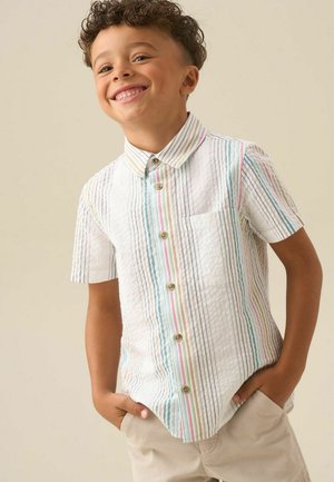 Ung dreng smiler, iført en kortærmet hvid skjorte med farverige lodrette striber og beige shorts, hænderne i lommerne.