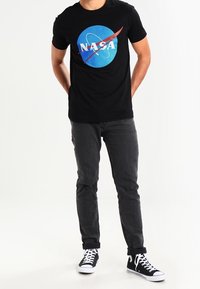 Svart bomullströja med blå NASA-logotyp i cirkulär design, kombinerad med mörkgrå jeans och svart-vita sneakers.