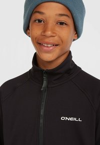 Zwarte zip-up jas met hoge kraag, voorzien van het witte O'Neill-logo. Draagt een lichtblauwe gebreide beanie, met een gladde textuur en aansluitend ontwerp.