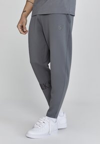 Pantalones de chándal grises de corte holgado, con bolsillos laterales y un logo bordado en el muslo. Combinados con zapatillas deportivas blancas.