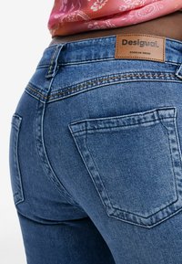 Jeans i denim med midjhöjd, kopparstickning och en läderlogga. Nära bild av bakfickorna och den släta texturen.