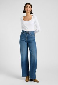 Top bianco a maniche lunghe con scollo quadrato abbinato a jeans blu a vita alta e gamba ampia, con stivali tan a tacco largo.