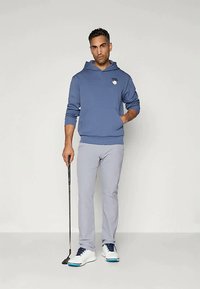 Άνδρας με μπλε φούτερ και ανοιχτό γκρι παντελόνι κρατάει ένα γκολφ putter, στέκεται μπροστά σε ένα απλό ανοιχτό φόντο με το ένα χέρι στην τσέπη.