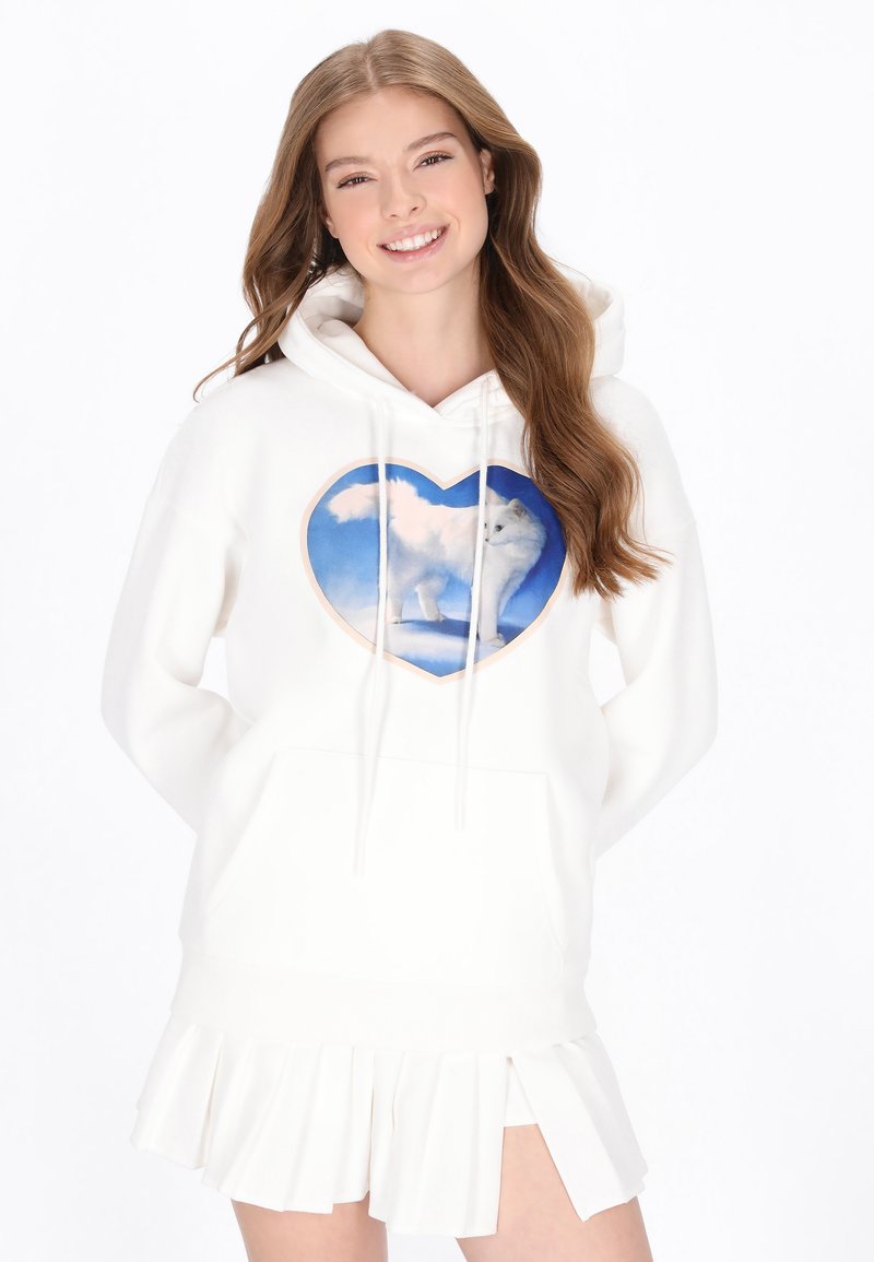 Fehér pulóver kapucnis felső szív alakú grafikával, amelyen egy fehér róka látható kék ég háttérrel, fehér pliszírozott szoknyával párosítva.