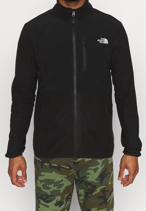 Czarna polarowa kurtka na zamek z kieszenią na piersi i logo The North Face, noszona z zielonymi i brązowymi spodniami w kamuflażu.