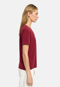 Czerwona koszulka z krótkim rękawem, zaprojektowana w dekoltem w serek, wykonana z miękkiego materiału, gładka w dotyku, luźno dopasowana, zestawiona z jasnymi spodniami.