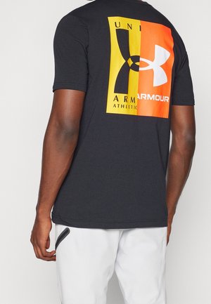 Person iført sort kortærmet skjorte med stort gult og orange Under Armour-logo på ryggen, kombineret med hvide bukser med sort lynlåsdetalje.