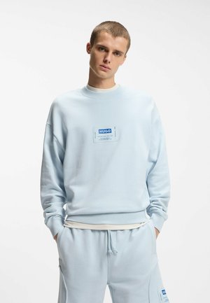 Młody mężczyzna w jasnoniebieskiej bluzie HUGO i pasujących spodniach dresowych, stojący z rękami w kieszeniach na jednolitym białym tle.