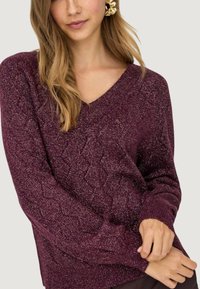 Maglione a maglia color bordeaux con scollo a V, caratterizzato da un motivo a diamante testurizzato e polsini a coste. Tessuto morbido con una leggera lucentezza.