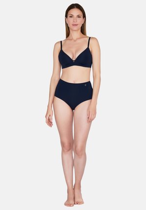 Kvinde står barfodet iført en navyblå bh med justerbare stropper og trusser med høj talje mod en ensfarvet hvid baggrund.
