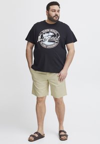 Svart grafisk t-skjorte med hvit tekst, khaki shorts og brune sandaler. T-skjorten har et rundt logo-design. Avslappet antrekk.